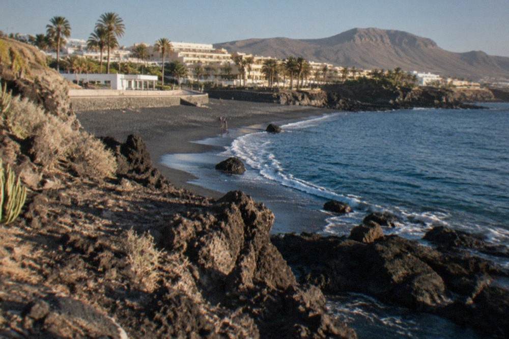 A quiet volcanic cove on the Tenerife coast with black sand and rock formations in the foreground