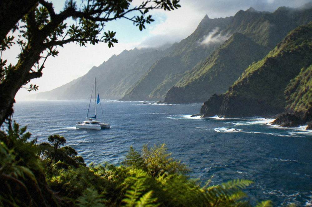 A catamaran with a blue flag sailing on open Atlantic water near a dark headland