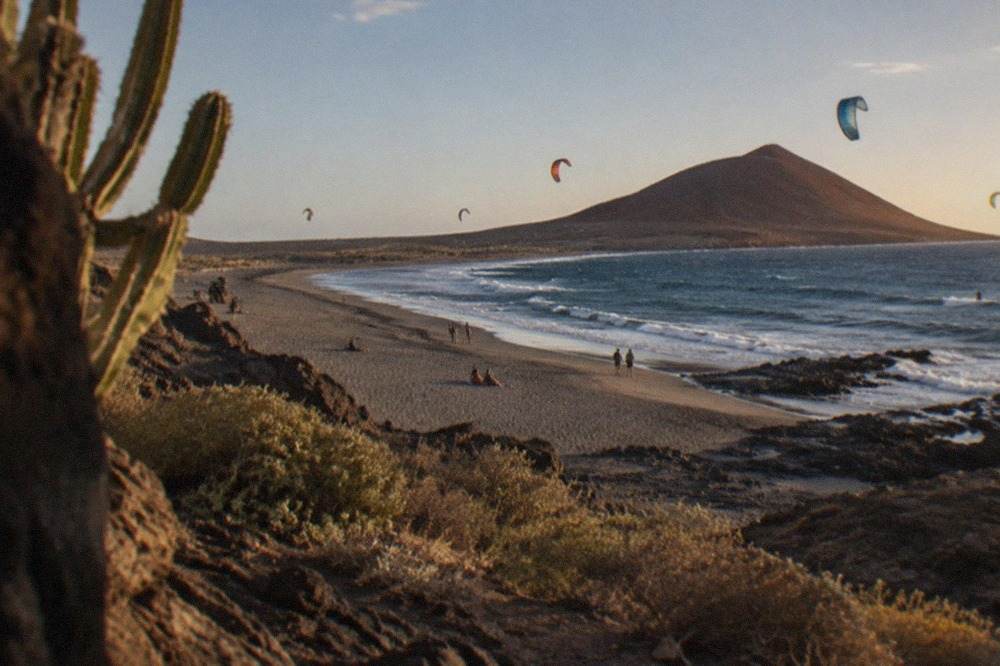A long grey sand beach curving along a bay with a red volcanic cone rising at its far end