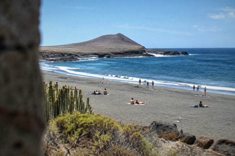 A wide bay of grey volcanic sand with sunbathers and a rocky point in the distance