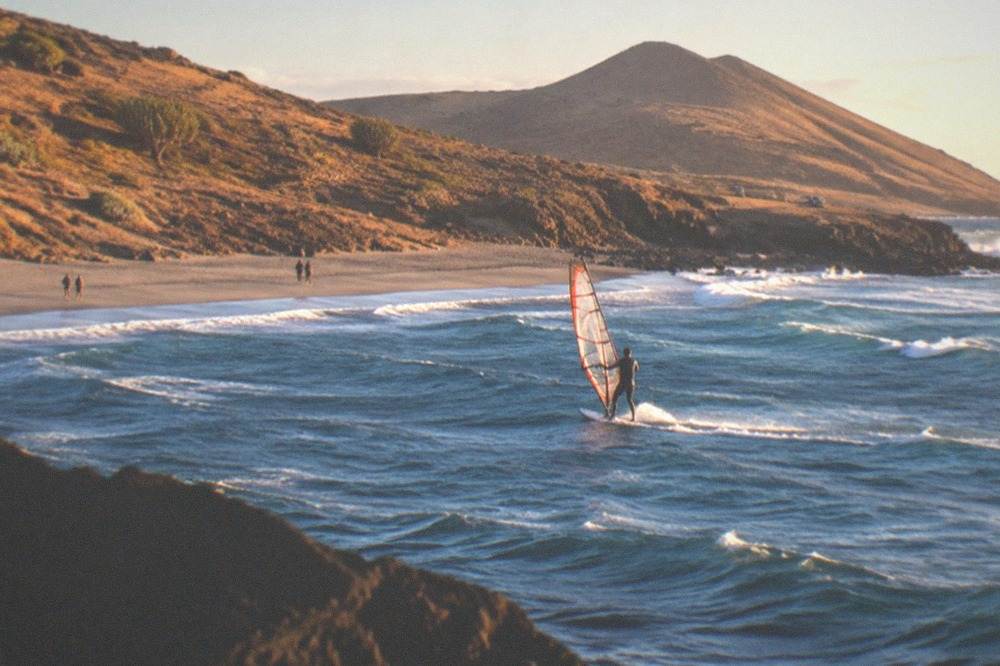 A windsurfer balancing on a board in a windy bay with a grey sand shore behind