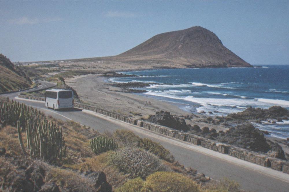 A coastal road beside a grey sand beach with a bus stop sign and palm trees