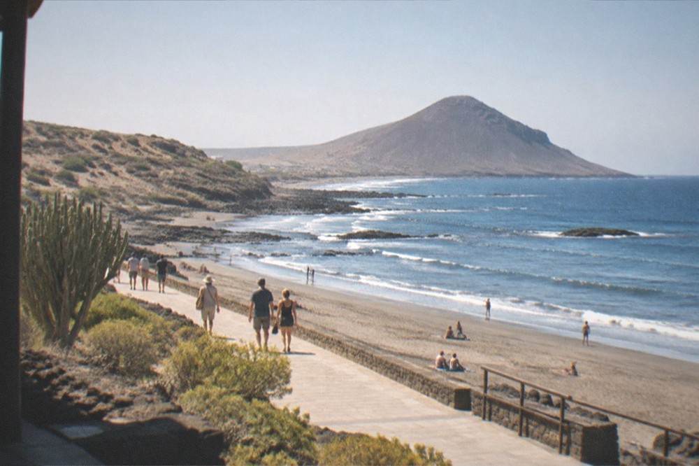 A sunny promenade along a grey sand bay with people walking and a calm sea beyond