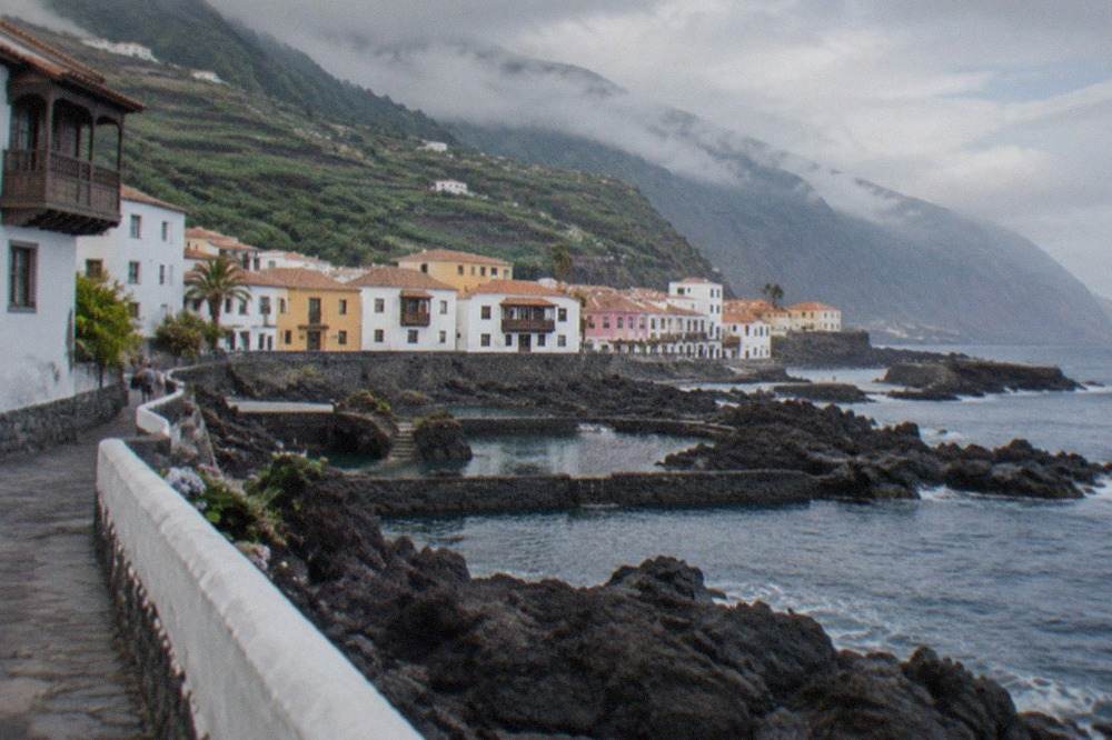 White and pastel houses of a small coastal town beside dark volcanic rock pools along the seafront