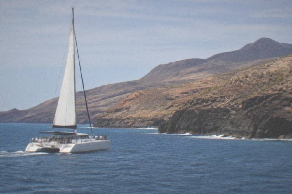 A catamaran with passengers sailing on open blue water near a volcanic coastline