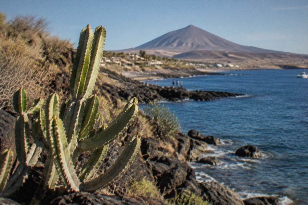 A rocky shoreline with clear water and a volcano cone in the distance under a blue sky