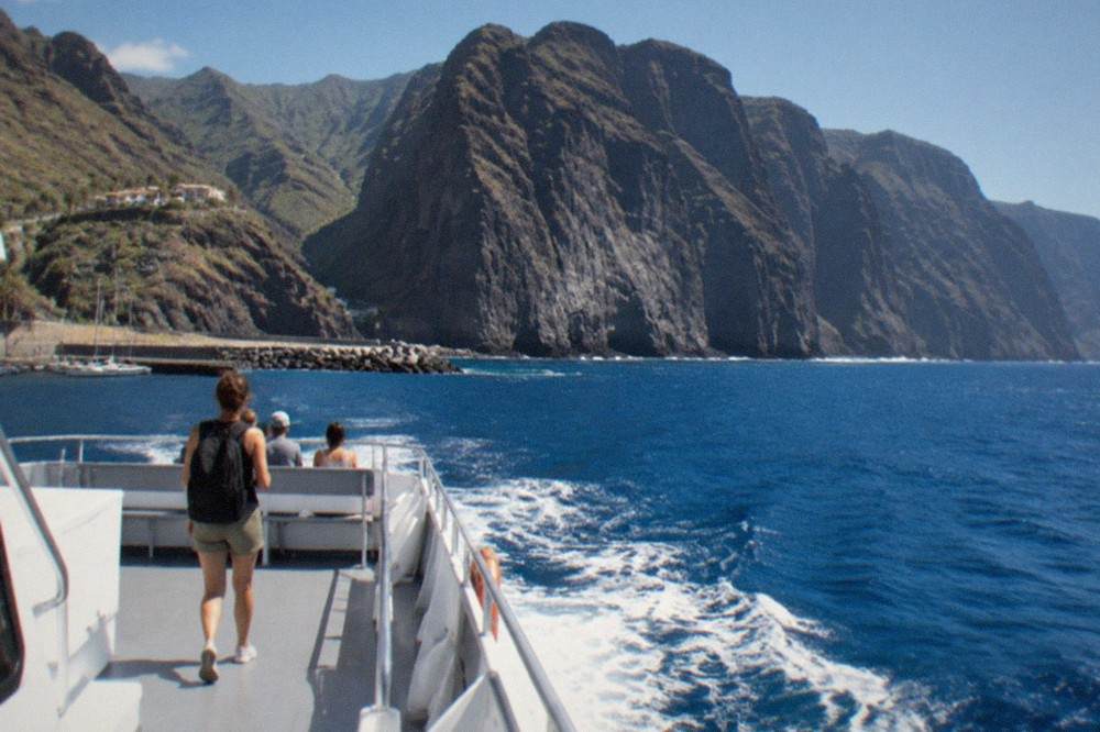 A passenger boat leaving a small marina towards tall dark sea cliffs in the morning