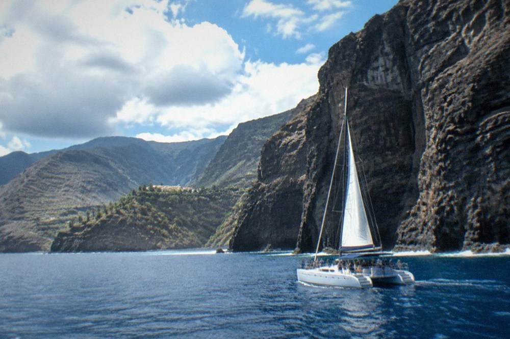 A catamaran with passengers sailing close to a tall dark cliff face above calm sea