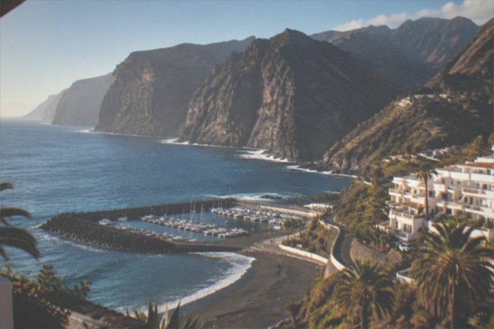 Low-rise white apartment buildings on a hillside above a small marina and dark sand beach