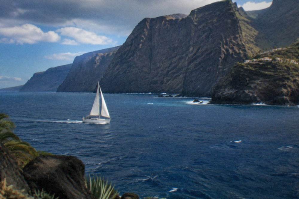 A white sailboat under way on open water with dark cliffs far behind