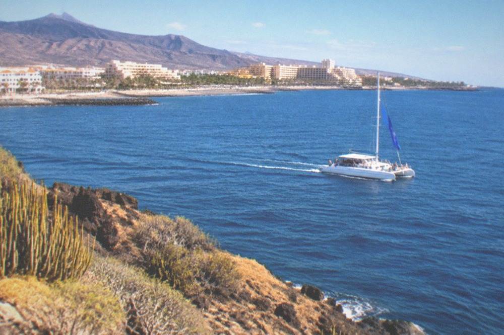 A catamaran under way on open blue water with a blue flag flying and passengers watching the sea