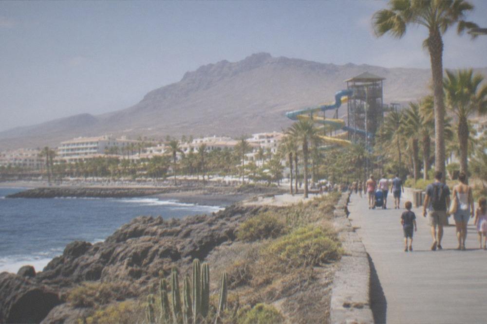 Families walking along a resort promenade with a water park slide tower visible behind palm trees
