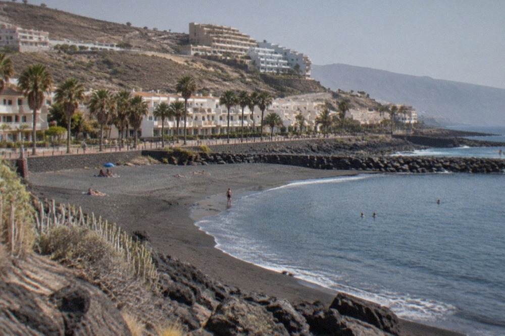 A calm swimming cove with dark sand, low sea walls and apartment blocks on the hillside behind