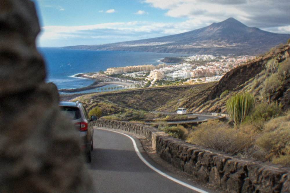 A winding mountain road descending towards green ravines and a distant volcanic peak