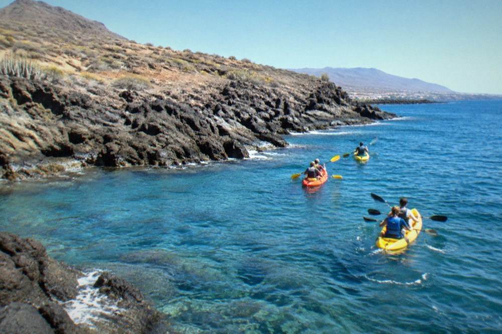 A kayak group paddling over clear water near dark rocks on the Tenerife coast