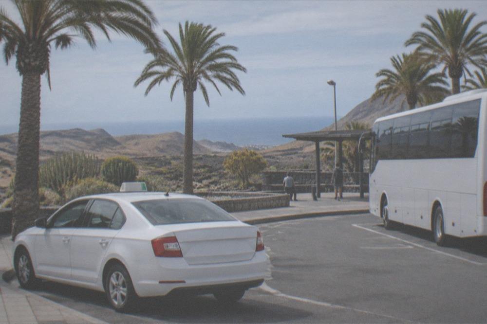 A white taxi and a bus at a small station forecourt under palm trees