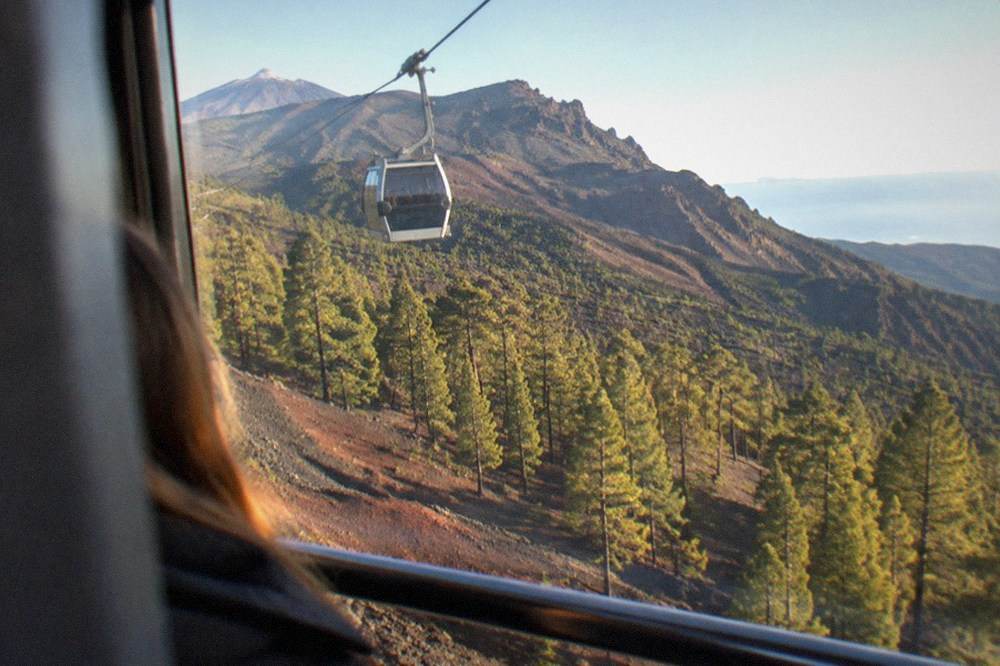 A cable car cabin travelling on cables above a red and grey volcanic mountainside