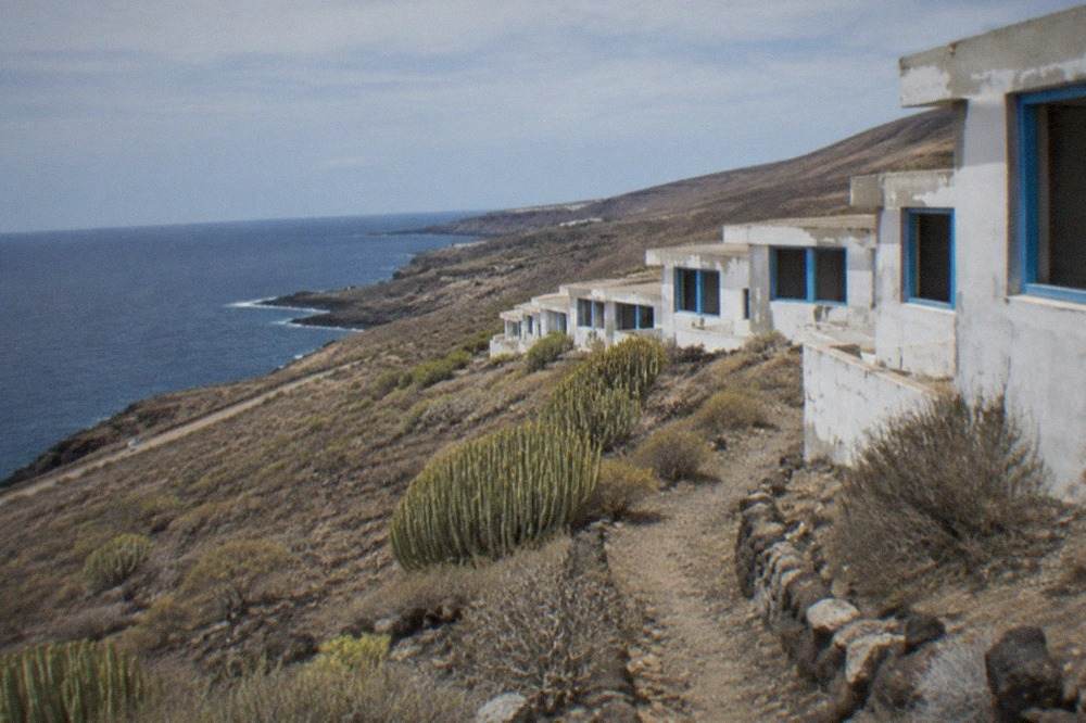 Rows of empty white abandoned buildings with blue window frames on a hillside above the sea