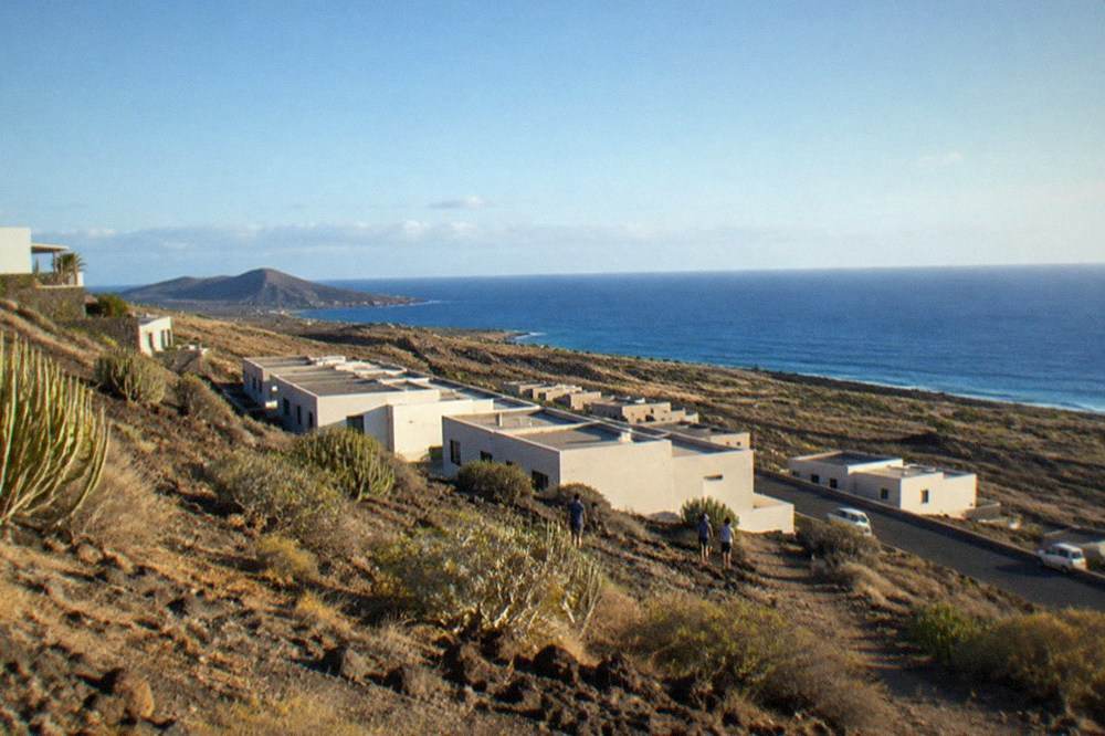 Empty rectangular buildings arranged in rows on a dry hillside with the sea in the distance