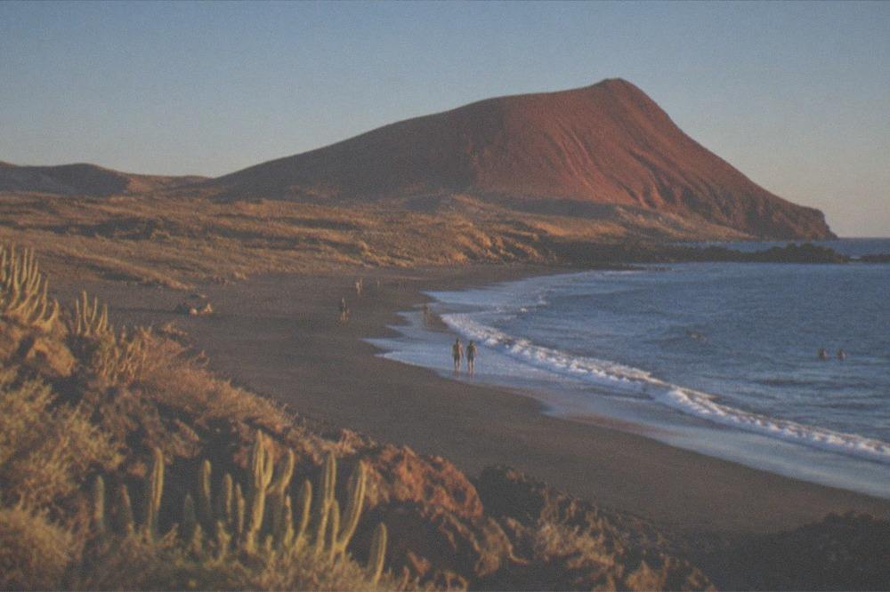 A wide sandy beach with a red mountain cone behind it under clear skies