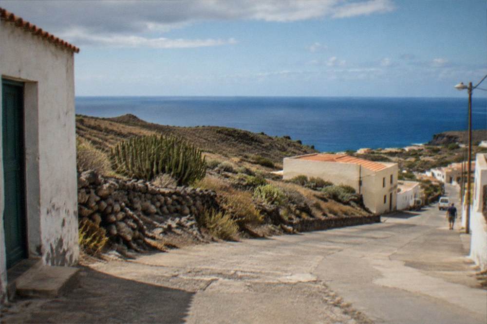 An empty village street with dry ground and the sea horizon beyond the rooftops