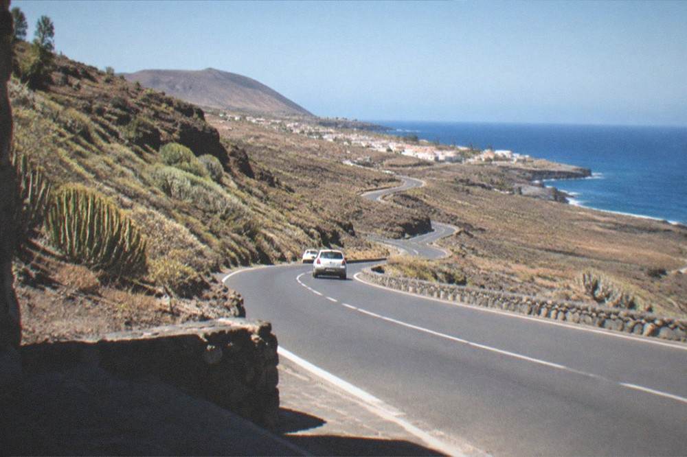 A coastal road winding past dry volcanic hills towards a distant village