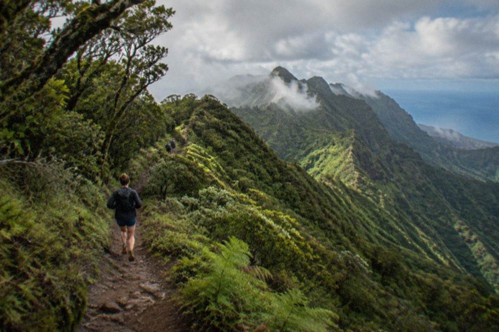 A narrow ridge trail winding between green slopes with cloud resting on distant peaks