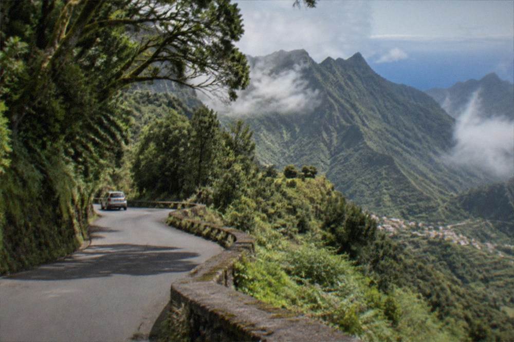 A narrow mountain road curving through green slopes with a distant village below