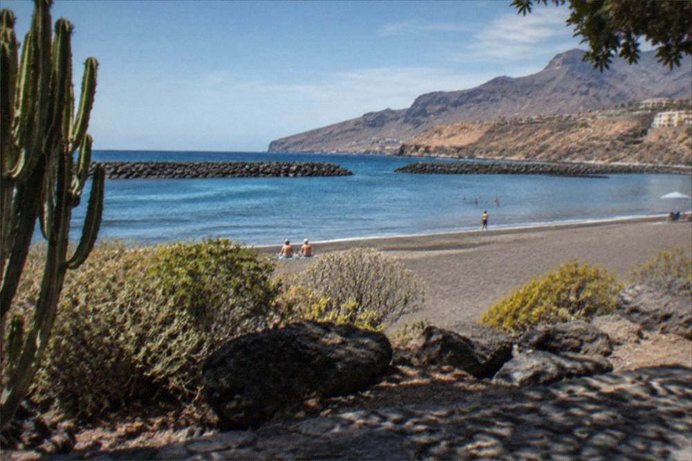 A sunny beach with grey sand and calm water behind breakwaters in Costa Adeje