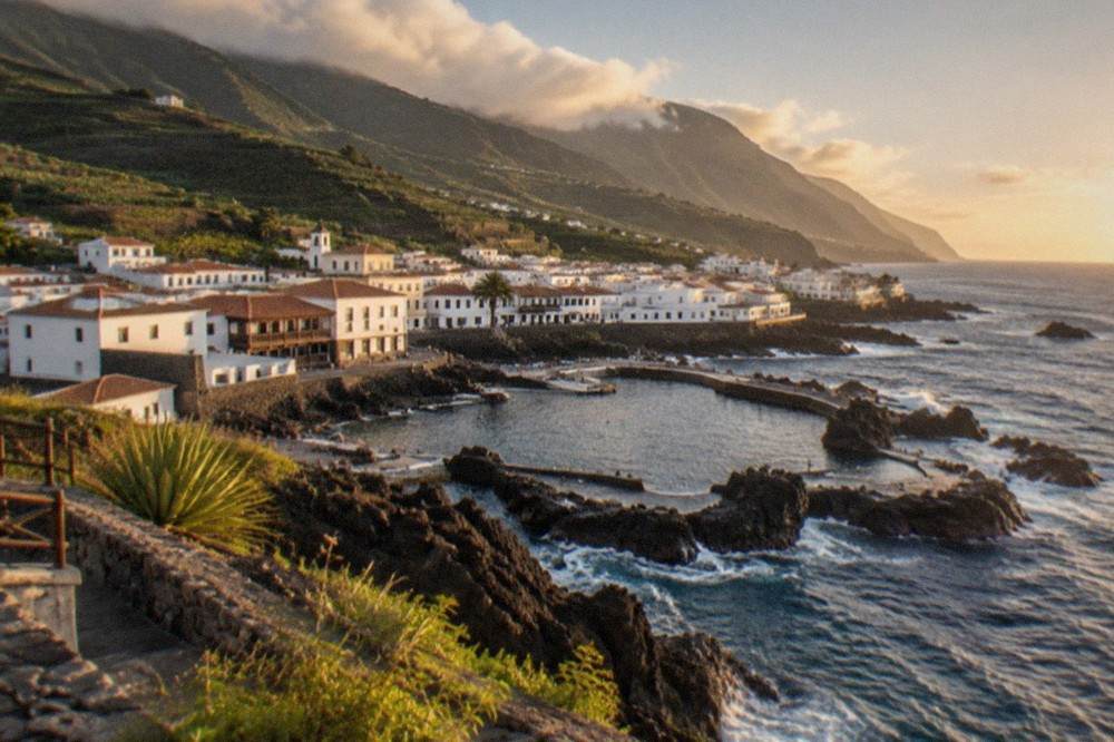 A wide view of Garachico town with low white buildings beside the sea and green hills behind