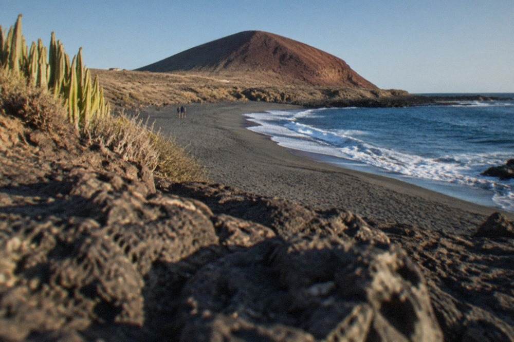 A long stretch of grey sand curving beneath a round red volcanic cone under a clear sky