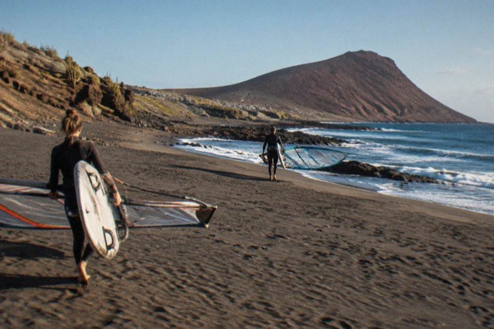 Wind surfers carrying boards across pale sand with a red volcanic cone in the background