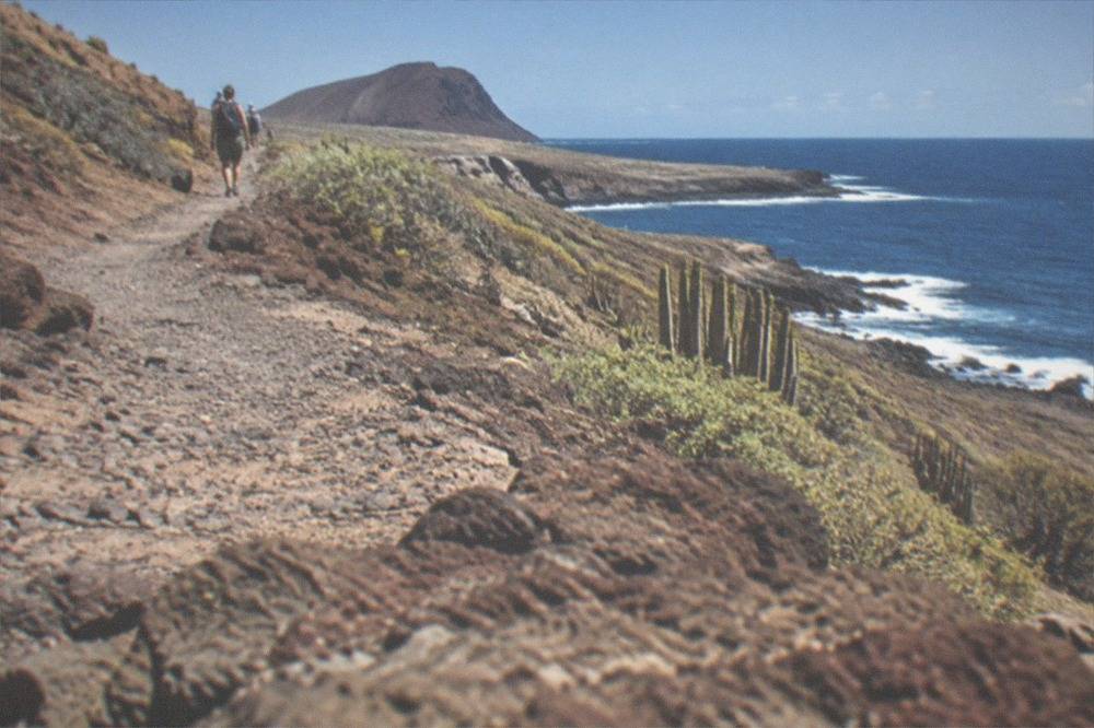 A gravel path rising over dark red volcanic rock with the coast visible below