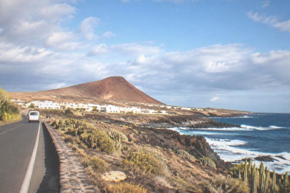 A coastal road with low white buildings and a red cone rising behind them