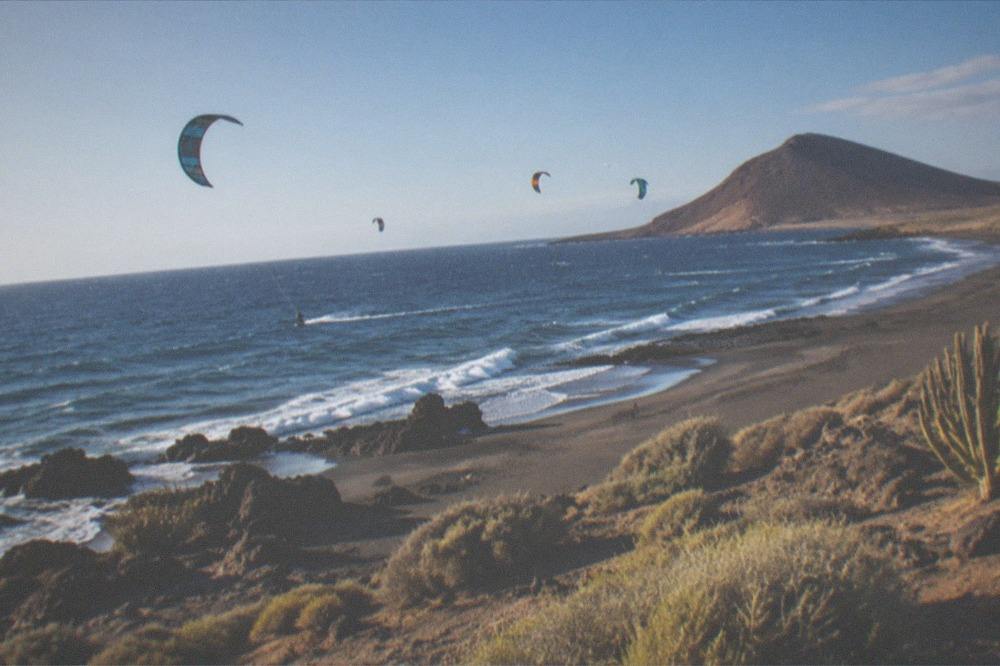 Kites flying over water with a wide sandy beach and scattered rocks below