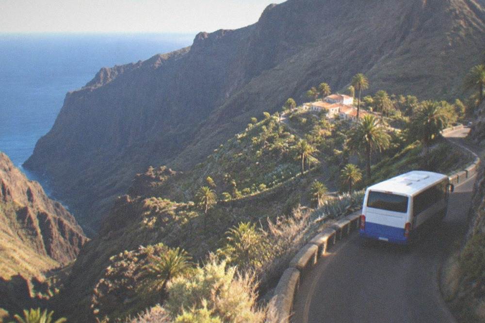A blue and white bus on a narrow mountain road with a deep valley to one side