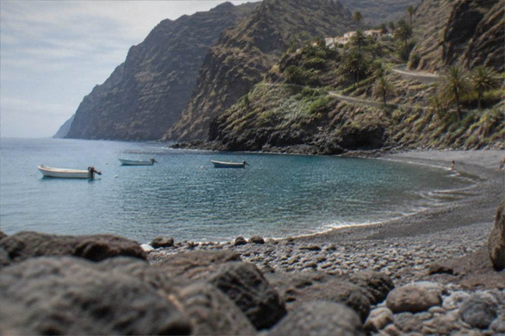 A small pebble beach at the foot of high dark cliffs with a few small boats on clear water