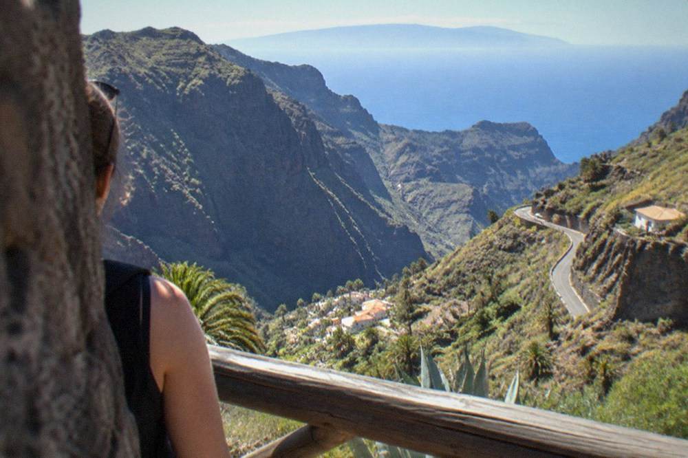 A coastal viewpoint above steep cliffs with a small village in a gorge below and a distant island on the horizon