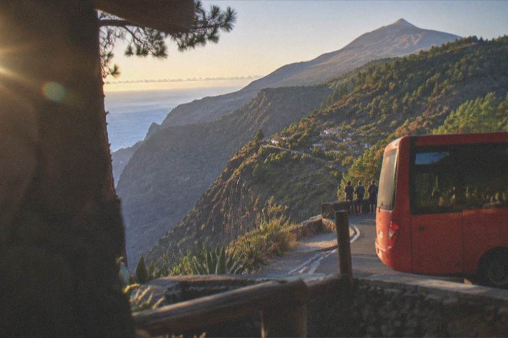 A red bus at a mountain stop with pine slopes and a volcanic peak behind it