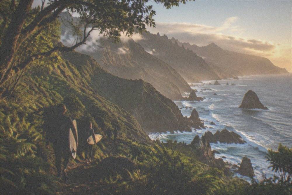 Surfers carrying boards along a path above a rocky Atlantic shoreline