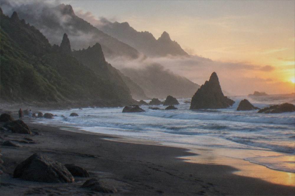 A quiet dark sand beach at dawn with waves and misty green mountains behind