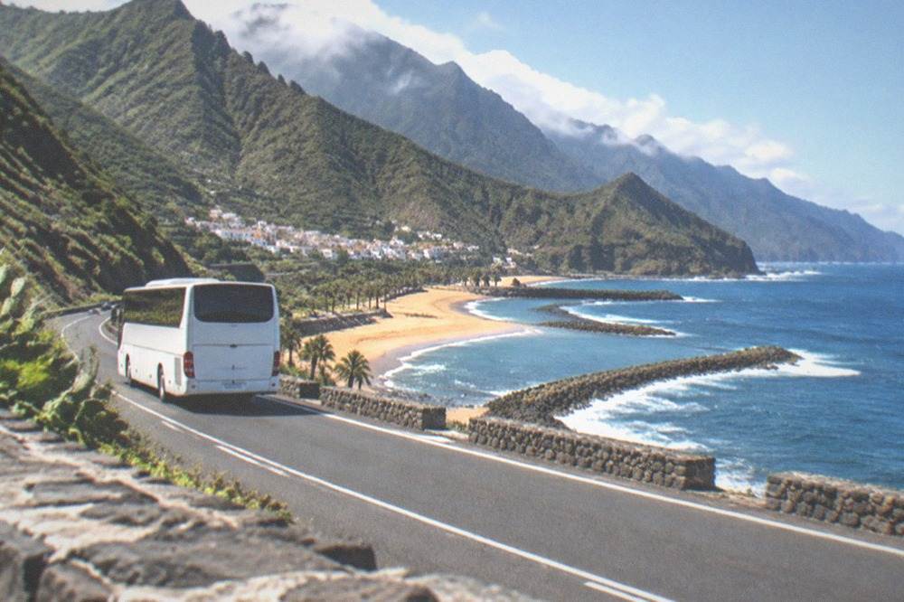 A white bus on a coastal road with a village and hills beyond