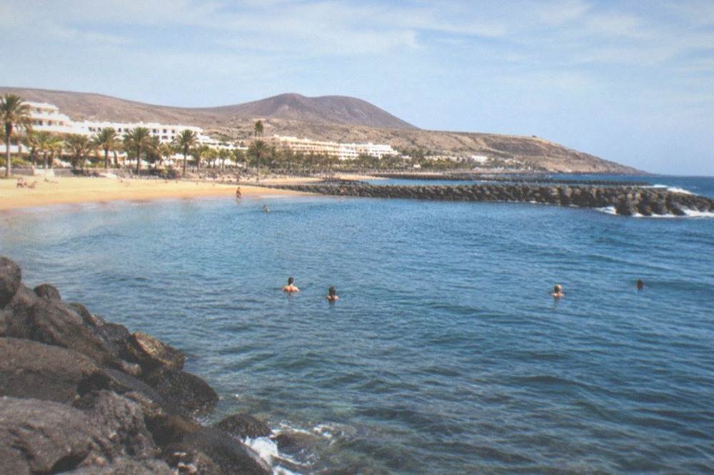 People swimming in calm clear water beside a stone breakwater on a sunny day
