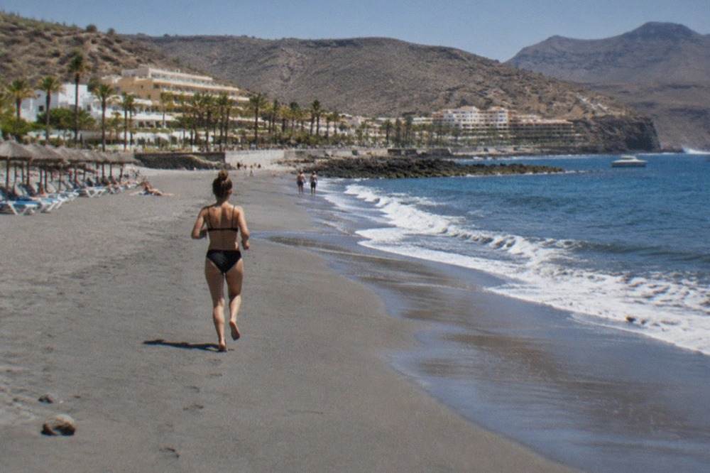 A wide grey-sand beach in front of hotels, with sun loungers and calm blue sea