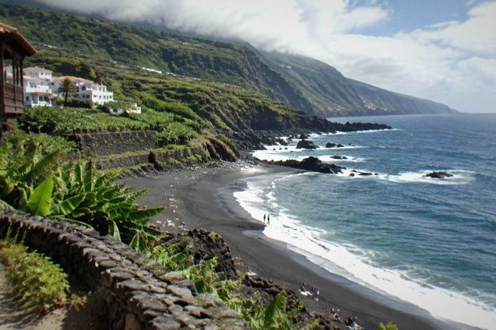 A stretch of dark sand curving below green cliffs under a cloudy sky