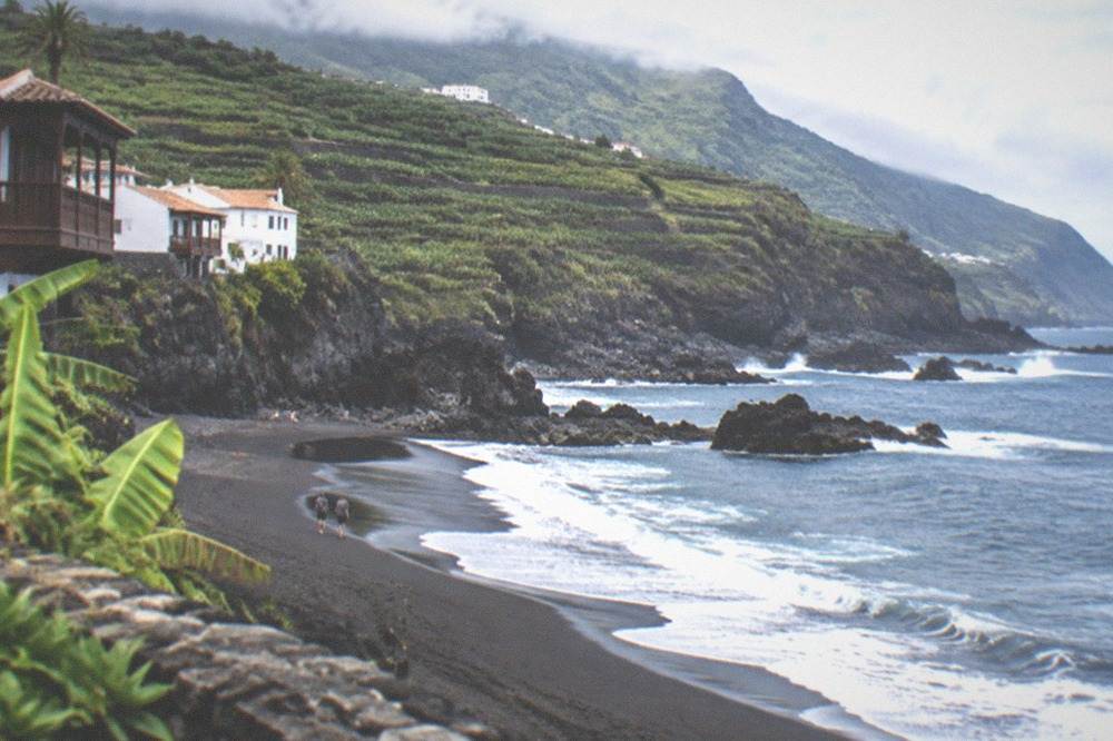 A dark sandy cove with surf and a green hillside under partial cloud
