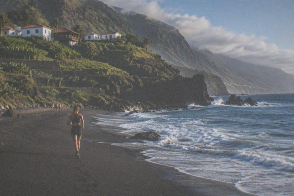 Dark sand meeting pale surf beneath a steep vegetated cliff