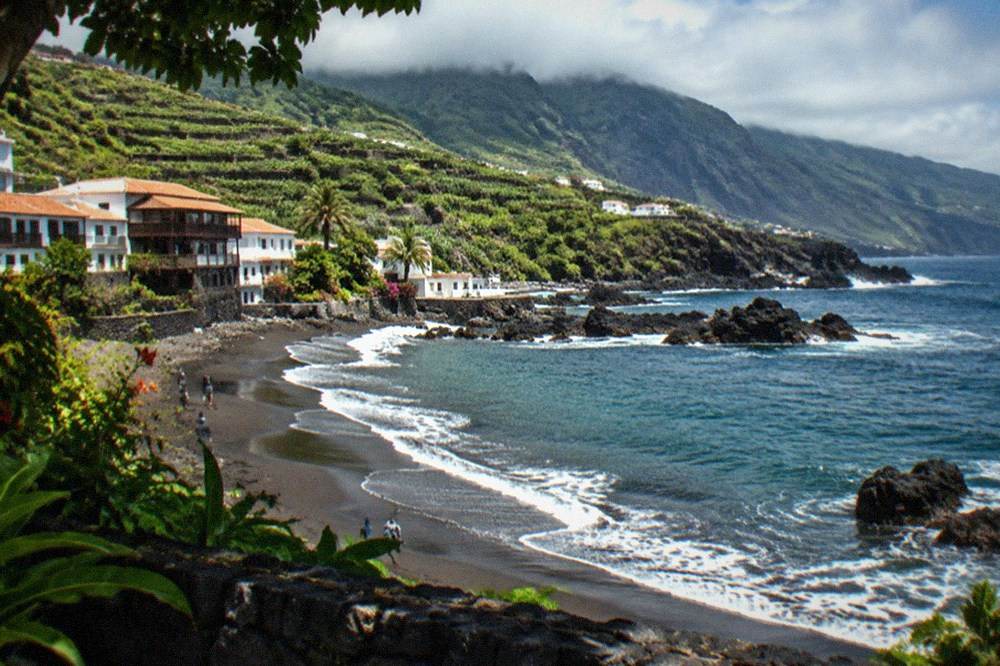 Dark volcanic sand meeting gentle surf along a garden-lined shore