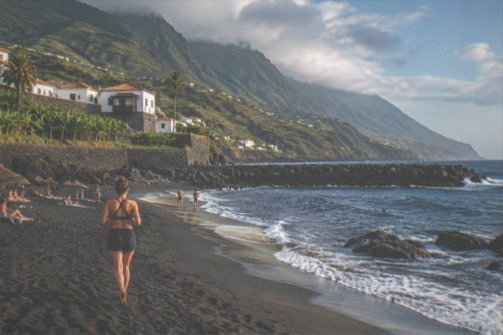 Sunbathers on dark sand with parasols and a stone breakwater in the sea