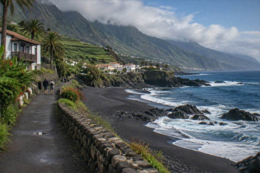 A coastal path with dark sand, gardens and distant mountains under afternoon light